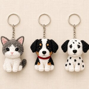 Puede incluir: Tres llaveros de ganchillo con figuras de animales: un gato gris y blanco, un perro negro, blanco y marrón con collar rojo, y un perro blanco con manchas negras. Cada figura está unida a una anilla y cadena de metal de tono plateado.