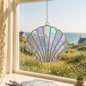 Puede incluir: Atrapasoles de concha de cristal manchado iridiscente colgado frente a una ventana. La concha está hecha de vidrio texturizado con un efecto arcoíris. La ventana da a un paisaje costero con un campo de flores silvestres y vistas al mar.