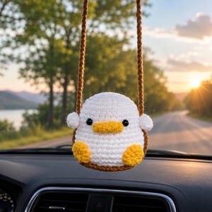 Puede incluir: Un adorno colgante de ganchillo con forma de pato blanco, pico amarillo y patas amarillas. El pato tiene pequeños ojos negros y alas blancas a los lados. Está suspendido por un cordón de ganchillo marrón para colgar del espejo retrovisor.