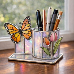 Puede incluir: Un portalápices decorativo con diseño de mariposa y tulipanes. El portalápices es de vidrio iridiscente y metal. La mariposa es naranja, amarilla y negra. Los tulipanes son rosas y naranjas. Varios bolígrafos están dentro.