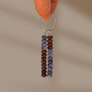 May include: A keyring featuring a silver-toned metal ring with two vertical strands of beads. One strand consists of blue and white mottled beads and brown beads, while the other strand features brown beads and blue and white mottled beads.