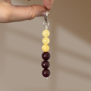 May include: A keyring featuring a silver-toned metal ring attached to a vertical strand of six spherical beads. The top three beads are a translucent pale yellow, while the bottom three beads are a deep, solid dark maroon colour.