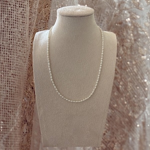 May include: A single strand necklace composed of small, uniform, off-white oval-shaped pearls. The necklace is displayed on a neutral-toned, fabric-covered bust form, showcasing the delicate arrangement of the individual pearl beads in a simple circular design.