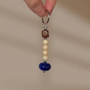 May include: A keyring featuring a silver metal ring attached to a vertical arrangement of beads. The sequence includes a single brown cube-shaped bead, three smooth cream-coloured spherical beads, and one large, glossy, deep blue disc-shaped bead.