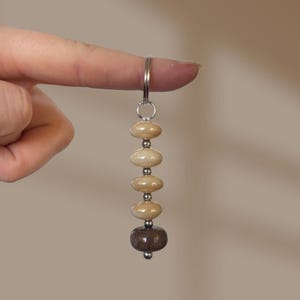 May include: A keyring featuring a silver metal ring attached to a vertical strand of beads. The strand consists of four smooth, light beige, oval-shaped beads separated by small silver spacers, ending with a single dark brown, round bead.