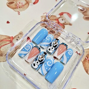 Puede incluir: Un juego de uñas postizas en una caja de plástico transparente. Las uñas son de color azul claro con diseños de remolinos blancos, acentos de copos de nieve y un muñeco de nieve de dibujos animados. Algunas uñas tienen una base nude con pedrería.
