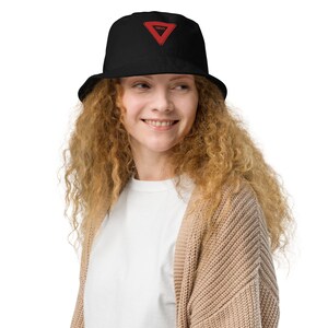 May include: A person wearing a black bucket hat with a red triangle and the word "YIELD" embroidered on it. The person is wearing a white t-shirt and a brown cardigan.