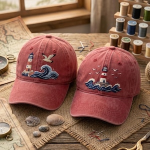 Op de afbeelding: Twee vervaagde rode baseballpetten met geborduurde nautische ontwerpen. Elke pet toont een vuurtoren, gestileerde blauwe oceaangolven en witte meeuwen. Het borduurwerk is gedetailleerd met witte, blauwe en oranje draad op de stof.