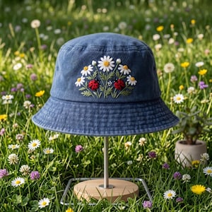 May include: A blue denim bucket hat featuring an embroidered design of white daisy flowers with yellow centers and two red ladybugs with black spots. The embroidery is centered on the front panel of the hat, showcasing detailed green stems and leaves.