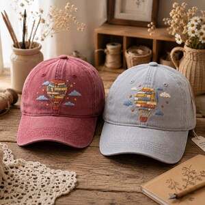 May include: Two baseball caps, one in a faded red and one in a light blue, each featuring an embroidered design of a hot air balloon constructed from a stack of colorful books, surrounded by small embroidered clouds and yellow stars.