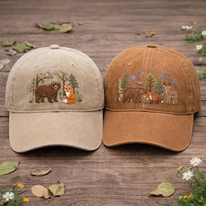 Op de afbeelding: Twee baseballpetten met geborduurde bosscènes. De ene pet is beige met een beer en een vos, de andere is bruin met een beer, een vos, een hert en vogels. Beide petten hebben een gebogen klep en een verstelbare band.