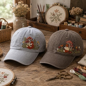 May include: Two baseball caps, one light gray and one dark brown, each featuring an embroidered design of a small orange fox sitting among red-capped mushrooms, green grass, and tiny colorful flowers on the front panel of the crown.