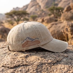 Puede incluir: Una gorra de béisbol de color beige con un diseño bordado en el lateral. El bordado muestra una cordillera con contornos en azul y terracota, junto a un pequeño sol amarillo sobre los picos. La gorra tiene visera curva y ojales.