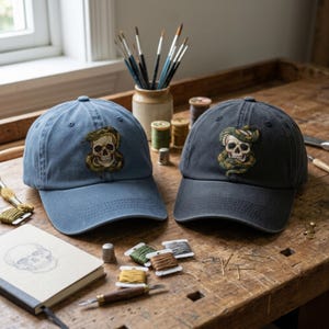 May include: Two baseball caps, one in dusty blue and one in charcoal grey, each featuring an embroidered patch of a human skull entwined with a green snake. The caps are positioned on a wooden surface alongside various sewing supplies and tools.