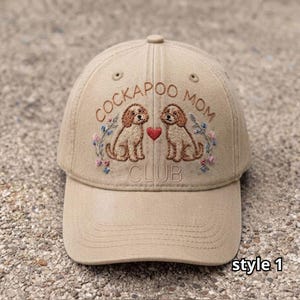 Puede incluir: Una gorra de béisbol de color tostado con el texto bordado COCKAPOO MOM CLUB. El diseño incluye dos perros marrones sentados a cada lado de un pequeño corazón rojo, rodeados de delicados bordados florales en tonos azules y rosas.