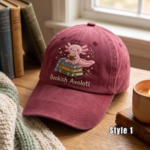 May include: A maroon baseball cap featuring an embroidered design of a pink axolotl sitting on a stack of books. The text "Bookish Axolotl" is embroidered below the illustration. The cap has a curved brim and stitched eyelets on the crown.