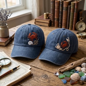 May include: Two blue denim baseball caps featuring embroidered designs. The left cap displays a red crab and a white seashell. The right cap features a red crab, a white seashell, and a rope detail. Both caps have metal eyelets on the crown.
