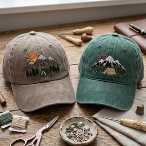 Puede incluir: Dos gorras de béisbol, una de color beige y otra verde, cada una con un bordado de un paisaje montañoso con sol, picos nevados, pinos y una pequeña tienda de campaña. Las gorras tienen una corona estructurada y una visera curva.