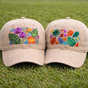 Puede incluir: Dos gorras de béisbol beige con diseños bordados de flores y hojas. Una gorra presenta orquídeas moradas y hojas verdes, mientras que la otra muestra hojas coloridas y formas geométricas. Ambas gorras están sobre una superficie de hierba verde.