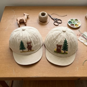 May include: Two beige baseball caps featuring embroidered designs of a brown bear standing next to a green pine tree. The caps are constructed from a light-colored fabric with a structured crown, a curved brim, and a small button at the top center.