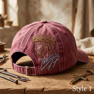 Peut inclure: Une casquette de baseball rouge délavé avec une broderie représentant le profil d'une figure barbue portant une couronne d'épines. Le motif est cousu en fil doré et bleu clair. La casquette possède une sangle réglable avec une boucle en métal.