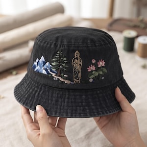 May include: A black bucket hat featuring detailed embroidery of blue snow-capped mountains, a green pine tree, a golden standing Buddha figure, and pink lotus flowers with green leaves arranged in a horizontal landscape design across the front.