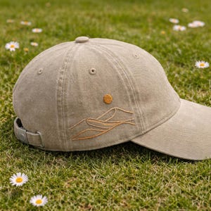 Puede incluir: Una gorra de béisbol de color beige con un diseño bordado de un sol y una cordillera en el lateral. La gorra cuenta con una corona estructurada, una visera curva y una correa ajustable con hebilla de metal en la parte posterior.