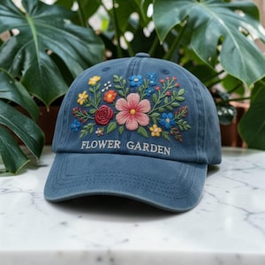 Puede incluir: Gorra de béisbol azul vaquero con un diseño floral bordado. El bordado presenta una variedad de flores coloridas, incluyendo rosas, rojas, amarillas y azules. El texto "FLOWER GARDEN" está bordado debajo.