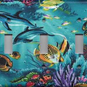 May include: A decorative triple light switch cover featuring an underwater scene. The design includes blue dolphins, colorful fish, a sea turtle, and coral against a teal background. The cover has three rectangular cutouts for the light switches.