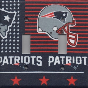 May include: A double light switch cover featuring a red, white, and blue American flag design with a New England Patriots football helmet and the words "Patriots Patriots".