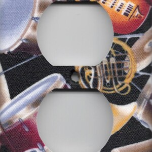 May include: A decorative light switch cover featuring a musical instrument design. The cover has a black background with images of a guitar, drums, and a French horn. The light switch openings are white.