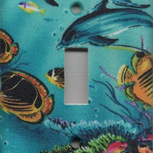 May include: A decorative light switch cover featuring an underwater scene. The design includes blue dolphins, colorful fish, coral, and aquatic plants against a teal background. The switch opening is a neutral gray color.