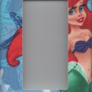May include: A light switch cover featuring a colourful illustration of Ariel, the Little Mermaid, with a blue, green and white background.