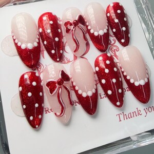 May include: A set of ten artificial nails with a red and white design. Some nails feature red glitter with white polka dots, while others have a light pink base with a white pearl border and a red bow design. The nails are oval-shaped.