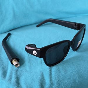 May include: Black sunglasses with a hidden compartment in the temple arm, resting on a blue surface. The sunglasses have dark lenses and a small logo on the frame. The temple arm is detached, revealing a rolled-up item inside.