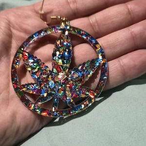 May include: A colorful, glitter-filled earring featuring a cannabis leaf design. The earring is a hoop shape with a gold-colored hook. The leaf is filled with a variety of colors, including blue, red, and green.