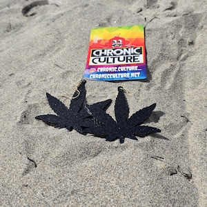May include: Black glitter leaf earrings with gold-colored hooks rest on sand. A colorful card with the text "CHRONIC CULTURE" and social media handles is also on the sand.