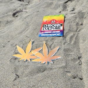 May include: Two leaf-shaped earrings with gold-colored hooks, featuring a gradient of orange and yellow. A colorful business card with the text "CHRONIC CULTURE" and social media handles is also visible. The items are displayed on a sandy surface.