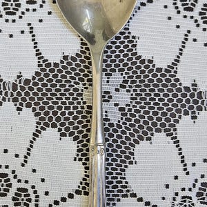 May include: A silver spoon with an oval bowl and a decorative handle. The handle has a detailed design with vertical lines and ornate flourishes. The spoon is likely vintage or antique, and is set against a white lace tablecloth.