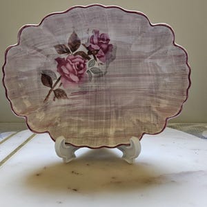 May include: A decorative plate with a scalloped edge and a floral rose design. The plate has a light purple background and a darker purple border. The roses are pink with brown leaves. The plate is displayed on a white stand.