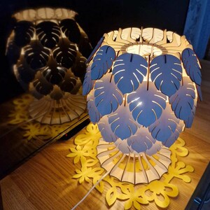 Puede incluir: Lámpara decorativa de madera con un diseño único. La lámpara presenta recortes en forma de hojas de color azul claro alrededor de la pantalla, con una base a juego. La lámpara está sobre una mesa de madera con un diseño floral amarillo.