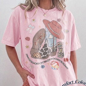 May include: A light pink t-shirt featuring a graphic print of a cowboy boot with a cow print pattern, a red cowboy hat, and stacked blocks with the letters T, S, and S. The design includes small flowers, a bow, and text reading I KNEW YOU.