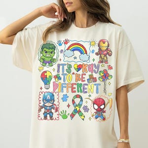 K&ouml;nnte beinhalten: Cremefarbenes T-Shirt mit farbenfrohen Cartoon-Illustrationen von Superhelden und dem Schriftzug "It's Okay to Be Different". Das Design beinhaltet einen Regenbogen, Puzzleteile und ein Band, das Inklusion und Akzeptanz f&ouml;rdert.