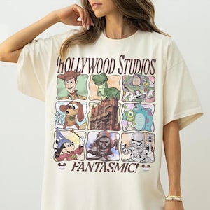 Può includere: T-shirt color crema con una griglia di personaggi dei cartoni animati e le parole "HOLLYWOOD STUDIOS" e "FANTASMIC!". La maglietta presenta personaggi come Woody, Buzz Lightyear e Mickey Mouse, con un'estetica vintage.
