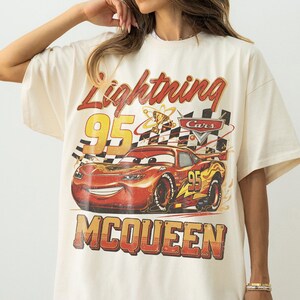 May include: Cream-colored t-shirt featuring a graphic of Lightning McQueen from the Cars movie. The design includes the text "Lightning McQueen 95" in red and yellow, with a checkered flag and a trophy.