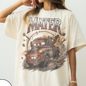 K&ouml;nnte beinhalten: Elfenbeinfarbenes T-Shirt mit einer Grafik von Hook aus dem Film Cars. Das Design enth&auml;lt das Wort "MATER" &uuml;ber dem Truck, mit einem Hintergrund aus Werkzeugen und Blitzen. Das Shirt hat kurze &Auml;rmel und eine lockere Passform.