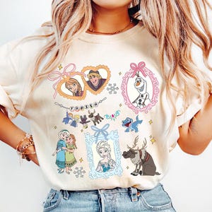 以下が含まれることがあります： アニメ映画「アナと雪の女王」のキャラクターのカラフルなイラストが描かれたクリーム色のTシャツ。デザインには、アナ、エルサ、クリストフ、オラフの額縁に入った肖像画、その他のキャラクター、そして「FROZEN」の文字が含まれています。