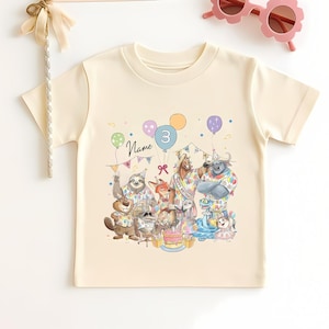May include: A cream-colored t-shirt featuring a colorful illustration of cartoon animals celebrating a birthday. The number 3 is on a balloon. Pink flower-shaped sunglasses and a banner that says "HOORAY" are also in the image.