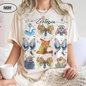 May include: An ivory t-shirt featuring the text Zootopia and a graphic print of various characters and bows. The design includes a blue snake, a fox, a buffalo, a beaver, a rabbit, and several colourful bows arranged around the central text.
