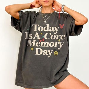 May include: Dark grey t-shirt featuring the text "Today Is A Core Memory Day" in white. The design includes stars, cherries, and a smiling face. The shirt has short sleeves and a loose fit.